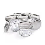 כמה מ-8 צנצנות זכוכית הברגה כפולה MASON JARS בנפח 118 מ"ל (4oz) עם מכסי מתכת מסודרים על רקע לבן, חלקם עומדים זקופים ואחרים מוטים או שוכבים.