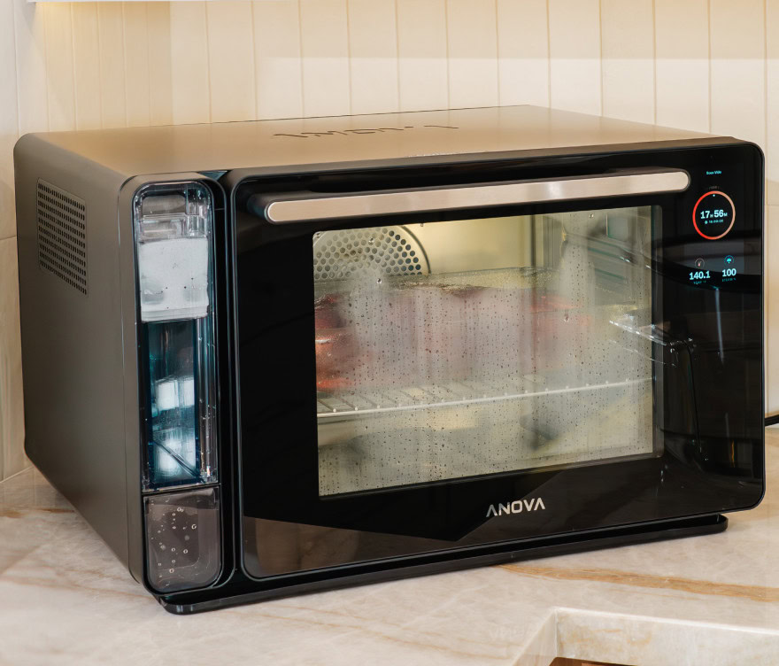 תנור Anova שולחני עם תצוגה דיגיטלית פועל, ומחמם את המזון שבתוכו; ניתן לראות אדים על דלת התנור.
