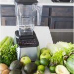 בלנדר של Blendtec מונח על השיש, מוקף בתפוחים ירוקים, אבוקדו, לימונים ירוקים, סלרי, חסה ועלים ירוקים אחרים.