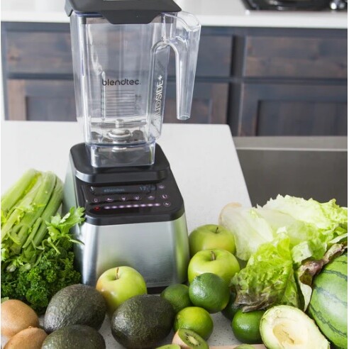 בלנדר של Blendtec מונח על השיש, מוקף בתפוחים ירוקים, אבוקדו, לימונים ירוקים, סלרי, חסה ועלים ירוקים אחרים.